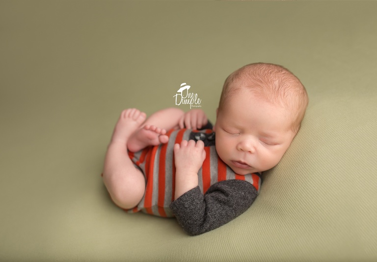 One Dimple Photography is a DFW, Southlake, Trophy Club, Grapevine, Argyle newborn photographer. In-Home Newborn Session baby stripped gray and orange outfit in back laying pose.